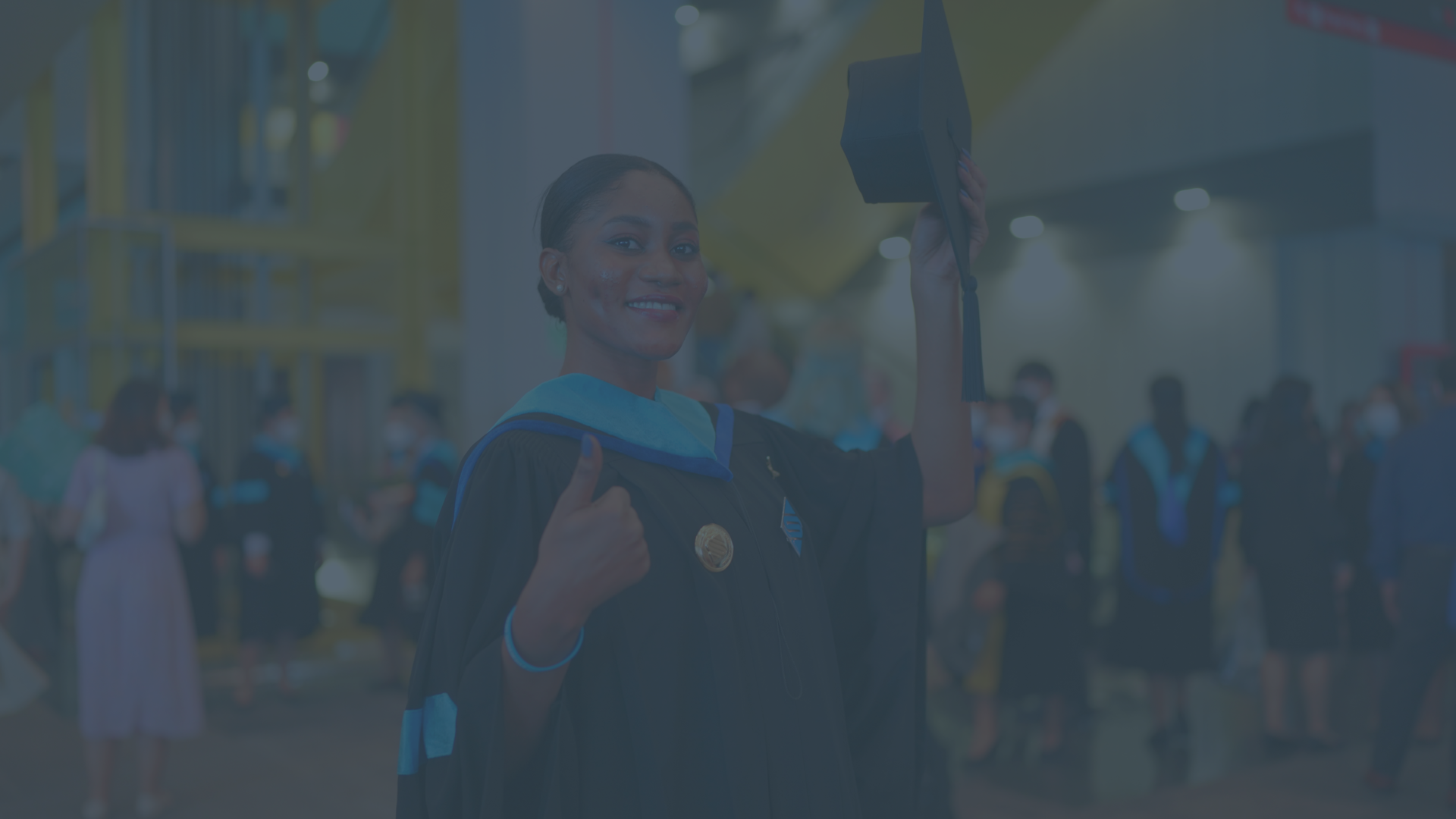 College graduate smiles at the camera and gives a thumbs-up sign with her right hand. There is a soft blue overlay.