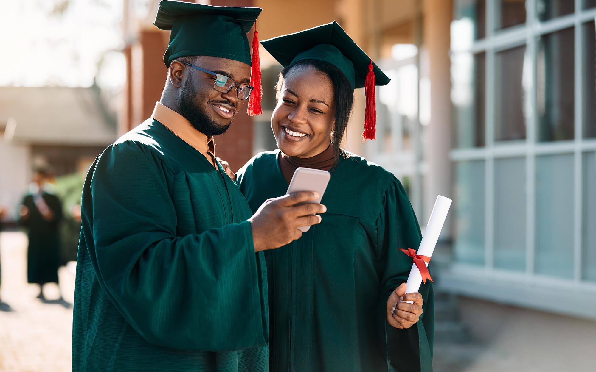 Two graduates holding diplomas and looking at photos on phone